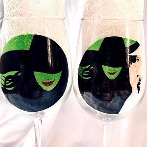 May include: Two wine glasses with hand-painted designs of the Wicked Witch of the West from the Wizard of Oz. The glasses feature a green face with a black hat and a black dress.