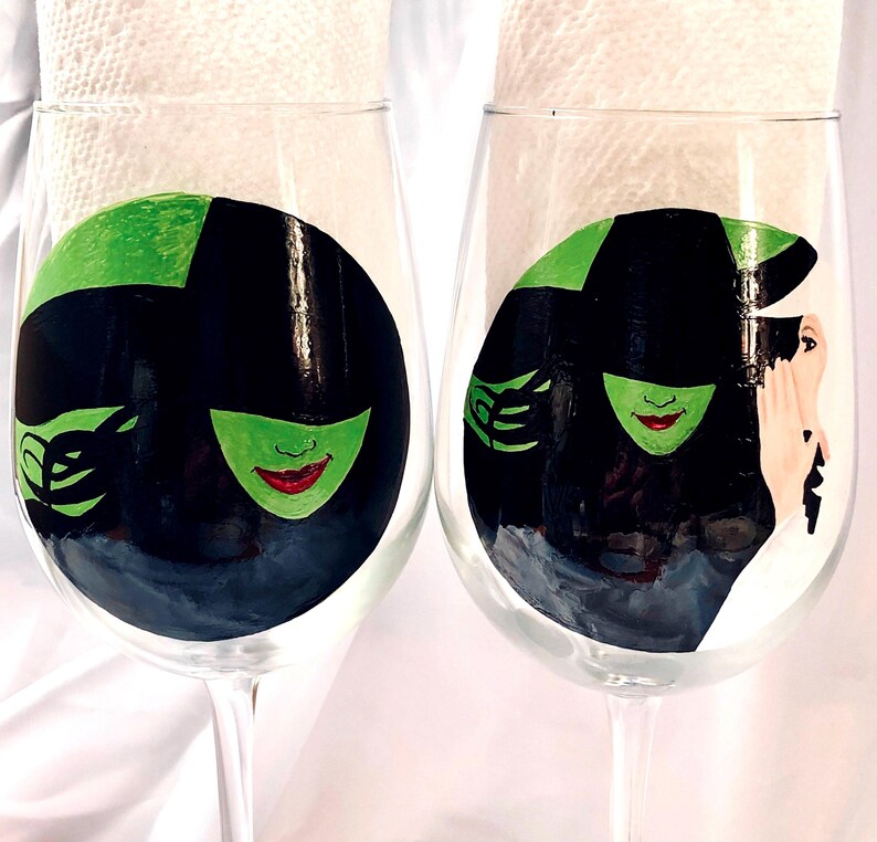 May include: Two wine glasses with hand-painted designs of the Wicked Witch of the West from the Wizard of Oz. The glasses feature a green face with a black hat and a black dress.