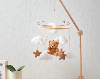 May include: A wooden baby mobile featuring a plush teddy bear, white clouds, brown stars, and a crescent moon. The mobile is suspended from a wooden arm and base, creating a whimsical nursery decoration.