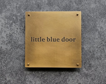 May include: A square, brass-colored sign with the words "little blue door" engraved in the center. The sign is mounted on a textured gray surface with four small brass-colored rivets in each corner.
