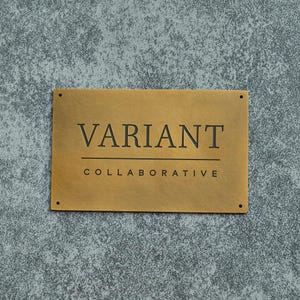 May include: A rectangular brass sign with the word "VARIANT" above "COLLABORATIVE" in black, sans-serif font. The sign has four small holes, one in each corner. The background is a textured gray surface.