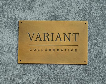 May include: A rectangular brass sign with the word "VARIANT" above "COLLABORATIVE" in black, sans-serif font. The sign has four small holes, one in each corner. The background is a textured gray surface.