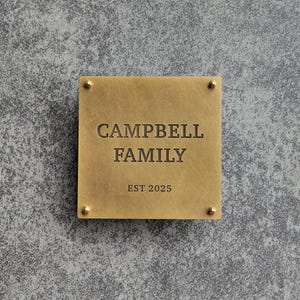Puede incluir: Placa cuadrada de color latón con el texto grabado "CAMPBELL FAMILY" y "EST 2025". La placa está asegurada con cuatro pequeños remaches dorados en cada esquina. El fondo es una superficie gris texturizada.