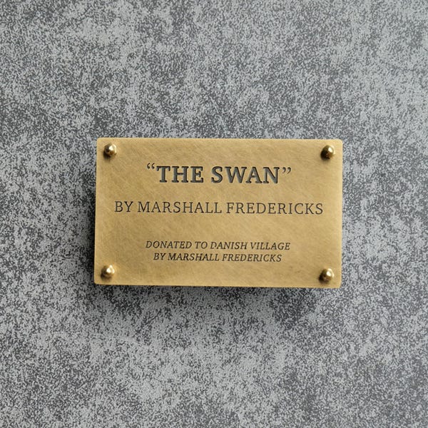 May include: A rectangular brass plaque with four rounded corner rivets. The text reads: "THE SWAN" BY MARSHALL FREDERICKS, DONATED TO DANISH VILLAGE BY MARSHALL FREDERICKS. The metal surface has a brushed finish and dark engraved lettering.