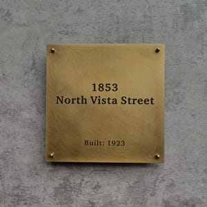 May include: A square brass address plaque with the address "1853 North Vista Street" and the text "Built: 1923" in black lettering. The plaque is mounted on a gray textured wall with four brass screws.