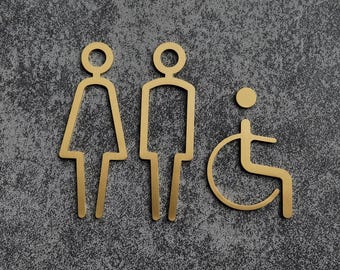May include: Gold-colored restroom signs for male, female, and accessible restrooms. The signs are minimalist in design, with simple line drawings of a person, a person in a wheelchair, and a female figure. The signs are on a gray textured background.