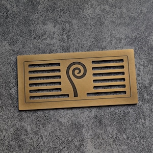 May include: A rectangular, bronze-colored metal vent cover with horizontal slits and a stylized fern design. The fern is in the center, with a spiral at the top and a curved stem extending downwards. The vent cover is on a gray surface.