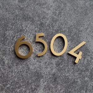 Pode incluir: Números de casa em metal dourado que formam o número "6504" contra um fundo cinza texturizado. Os números estão em uma fonte moderna e parecem ser peças individuais tridimensionais.