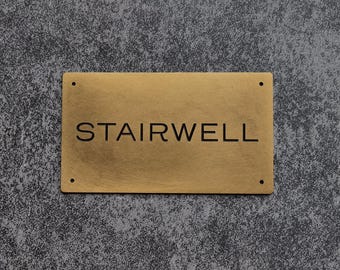 May include: A rectangular brass sign with the word "STAIRWELL" engraved in black, centered on the sign. The sign has four small holes, one in each corner. The background is a textured gray surface.