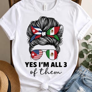 Peut inclure: Un t-shirt blanc à manches courtes avec un motif de chignon décoiffé portant un bandeau aux couleurs des drapeaux cubain, américain et mexicain. Le dessin inclut des lunettes d'aviateur aux verres drapés et le texte YES I'M ALL 3 of them.