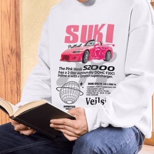 May include: White long-sleeve shirt featuring a pink graphic of a Honda S2000 sports car with the text "SUKI" and "Prettiest". The shirt also includes engine specifications and the text "Veilsi".