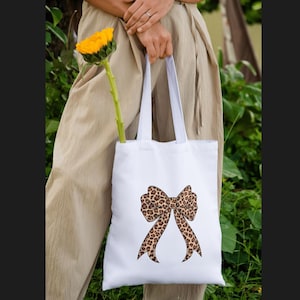 Può includere: Borsa tote bianca con un grande design a fiocco con stampa leopardata. La borsa è tenuta da una persona che indossa pantaloni beige. Un girasole giallo è posto all'interno della borsa.