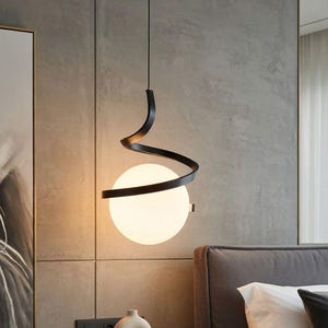 Może przedstawiać: Nowoczesna lampa wisząca z czarną, spiralną metalową ramą i kulistym, matowym szklanym kloszem. Lampa wisi nad stolikiem nocnym z małym wazonem, książkami i zegarem. Sypialnia ma neutralną paletę kolorów.