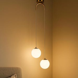 Op de afbeelding: Twee moderne hanglampen met bolvormige witte glazen kappen hangen aan een goudkleurige armatuur. De lampen hangen boven een nachtkastje met een fotolijst en een rietverspreider, waardoor een warme sfeer in een slaapkamer ontstaat.