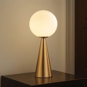 May include: A modern table lamp with a spherical, frosted glass shade. The lamp has a conical, brushed gold base. The lamp is turned on, emitting a warm, inviting glow. The lamp is placed on a dark wooden surface.