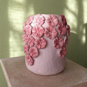 Op de afbeelding: Een cilindrische keramische vaas in een zachte pastelroze tint, voorzien van talrijke driedimensionale, met de hand gevormde roze bloemen die op het buitenoppervlak zijn aangebracht. De bloemen hebben gedetailleerde blaadjes en een glanzende afwerking.