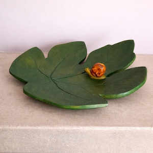 Op de afbeelding: Een groene bladvormige schaal met een kleine, getextureerde oranje en bruine slakfiguur bevestigd op het oppervlak. De schaal heeft zichtbare nerven die in het groene oppervlak zijn uitgesneden, wat zorgt voor een realistische botanische uitstraling.