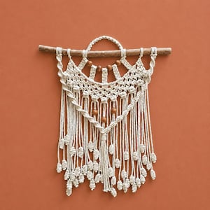 Pode incluir: Uma tapeçaria de macramé feita com corda de algodão em tom off-white, com nós num padrão decorativo e suspensa num ramo de madeira horizontal. O design inclui um aro central, cinco contas de madeira castanha e longas franjas pendentes.