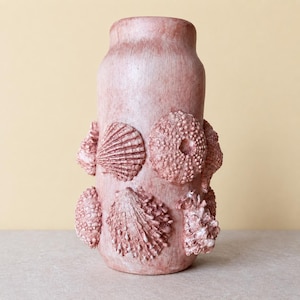 Pode incluir: Um vaso cilíndrico com acabamento mate em rosa empoeirado, decorado com vários elementos marinhos em relevo, incluindo conchas texturizadas e esqueletos de ouriços-do-mar dispostos em torno da superfície exterior do recipiente num padrão.