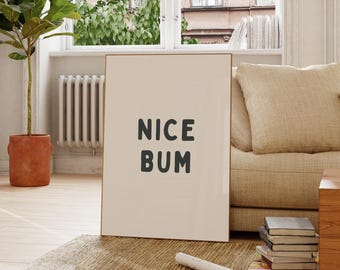 Op de afbeelding: Een ingelijste print met de woorden "NICE BUM" in donkergrijze letters. De print leunt tegen een beige bank in een kamer met een raam, een plant en een radiator.