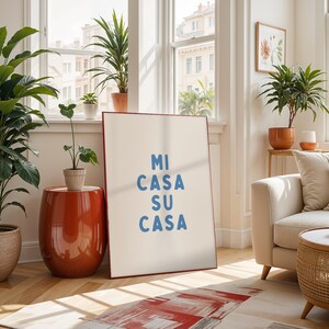 以下が含まれることがあります： 青い文字で「MI CASA SU CASA」と書かれた額入りのプリントがある、明るく風通しの良いリビングルームのシーン。 部屋には様々な鉢植えの植物、赤いサイドテーブル、ベージュのソファ、柄のラグがあり、居心地の良い雰囲気を作り出しています。