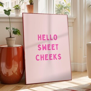 Peut inclure: Une affiche rose encadrée avec l'inscription "HELLO SWEET CHEEKS" en rose vif. L'œuvre est appuyée contre un mur dans une pièce lumineuse, à côté d'une table d'appoint orange brillante.