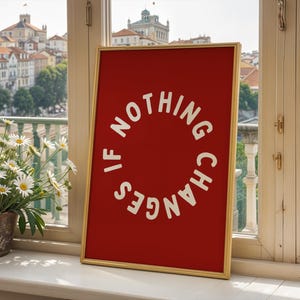 May include: A framed red art print with the text "IF NOTHING CHANGES" in a white circular design. The gold frame sits on a white windowsill, with a view of a European city and a pot of daisies.