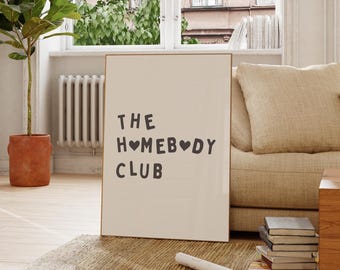 May include: A framed print with the text "THE HOMEBODY CLUB" in a black, sans-serif font. The word "HOMEBODY" features heart symbols. The print is displayed indoors, leaning against a wall, with a plant and sofa in the background.