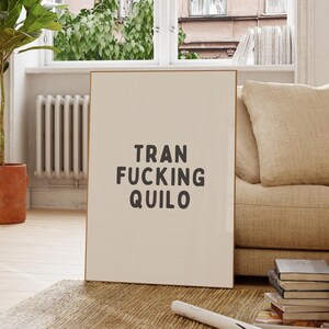 May include: A rectangular wall art print in a thin frame featuring a light beige background with the dark gray text TRAN FUCKING QUILO centered in a bold, sans-serif font. The print is displayed standing upright on a textured floor surface.