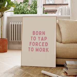 May include: A rectangular wall art print featuring a light beige background with the text BORN TO YAP FORCED TO WORK printed in a bold, pink, sans-serif font. The print is encased in a thin, metallic gold frame and stands upright on a textured surface.