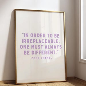 May include: A rectangular wall art print in a thin gold frame featuring the quote "In order to be irreplaceable, one must always be different." by Coco Chanel in light purple sans-serif lettering against a plain white background.