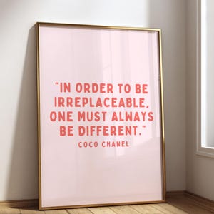Puede incluir: Un cuadro rectangular en un marco dorado fino con fondo rosa claro y texto de color coral que dice: "IN ORDER TO BE IRREPLACEABLE, ONE MUST ALWAYS BE DIFFERENT." seguido del nombre "COCO CHANEL" en un tamaño de letra más pequeño.