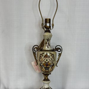 May include: A table lamp with a cream-colored ceramic base featuring a multi-colored floral and scrollwork pattern. The lamp has ornate brass-toned metal handles, a matching brass-toned base with feet, and a metal harp fixture at the top.