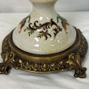 May include: A ceramic lamp base with a cream-colored crackle glaze finish, decorated with black, green, and brown scrollwork patterns. The base is supported by a circular, ornate, antique brass-toned metal stand featuring four decorative feet.
