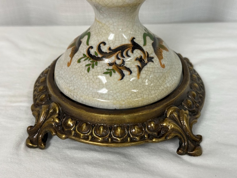 May include: A ceramic lamp base with a cream-colored crackle glaze finish, decorated with black, green, and brown scrollwork patterns. The base is supported by a circular, ornate, antique brass-toned metal stand featuring four decorative feet.
