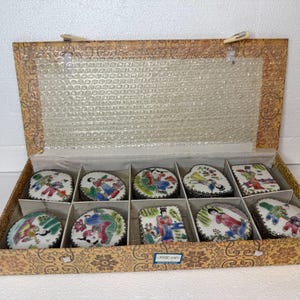 May include: A set of ten small, decorative porcelain boxes with lids, each featuring a unique hand-painted scene in vibrant colors. The boxes are arranged in a patterned cardboard box with a clear plastic cover.