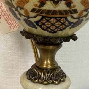 May include: A table lamp with a cream-colored ceramic base featuring a crackle glaze finish. The lamp has a brass-toned metal pedestal and a decorative ceramic body adorned with blue, orange, and black floral patterns and a warning tag.