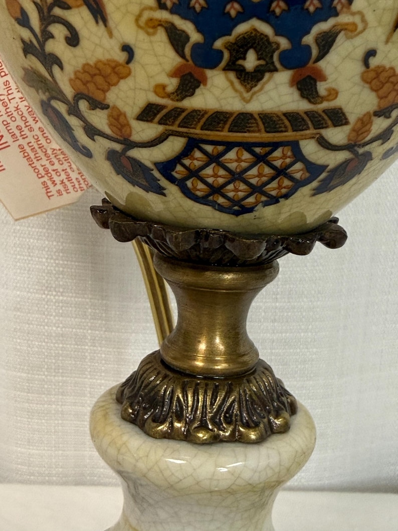 May include: A table lamp with a cream-colored ceramic base featuring a crackle glaze finish. The lamp has a brass-toned metal pedestal and a decorative ceramic body adorned with blue, orange, and black floral patterns and a warning tag.