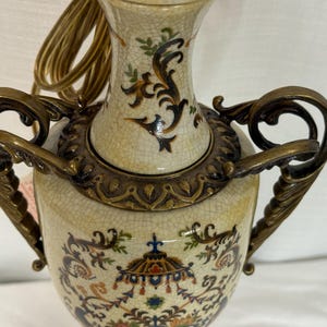 May include: A ceramic table lamp base with a cream-colored crackle glaze finish, featuring ornate bronze-toned metal handles and decorative accents. The body is adorned with intricate, multi-colored floral and architectural patterns.