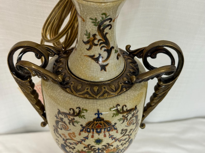 May include: A ceramic table lamp base with a cream-colored crackle glaze finish, featuring ornate bronze-toned metal handles and decorative accents. The body is adorned with intricate, multi-colored floral and architectural patterns.