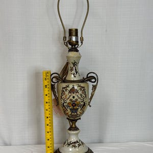 May include: A cream-colored ceramic table lamp with ornate brass handles and base, featuring a colorful floral and bird motif. A yellow measuring tape placed next to the lamp indicates a height of approximately 11 inches to the bulb socket.