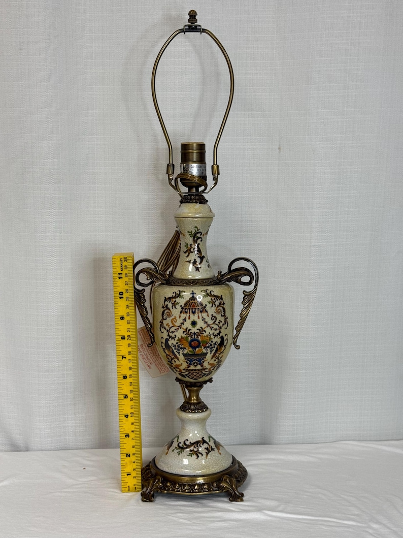 May include: A cream-colored ceramic table lamp with ornate brass handles and base, featuring a colorful floral and bird motif. A yellow measuring tape placed next to the lamp indicates a height of approximately 11 inches to the bulb socket.