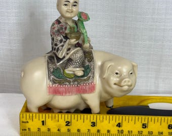 May include: A cream-colored figurine of a person riding a pig, positioned behind a yellow ruler measuring approximately 6 inches in length. The person wears a patterned robe and holds a green flower, while the pig stands on a flat surface.