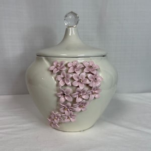 Puede incluir: Un tarro de cerámica blanca con tapa a juego rematada por un remate de cristal facetado transparente. El exterior presenta un grupo vertical de pequeñas flores esculpidas de color rosa claro con centros dorados en un patrón en cascada.