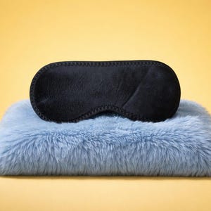 May include: A black sleep mask rests on a light blue, fluffy pillow. The sleep mask is oval-shaped with a stitched border. The pillow has a soft, furry texture. The background is a solid yellow.