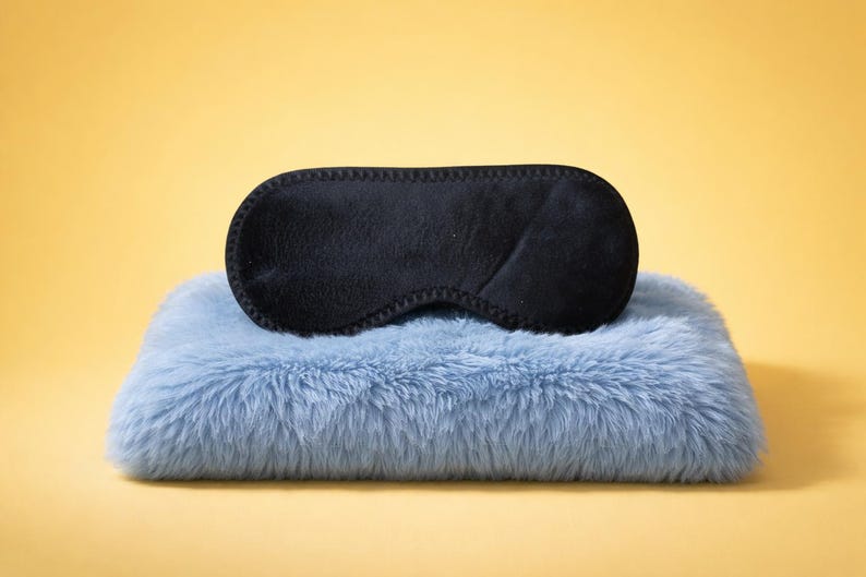 May include: A black sleep mask rests on a light blue, fluffy pillow. The sleep mask is oval-shaped with a stitched border. The pillow has a soft, furry texture. The background is a solid yellow.