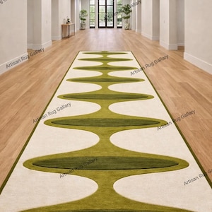 May include: A long, rectangular rug with an abstract design in a hallway. The rug features a cream-coloured background with olive green oval shapes. The rug is bordered with a thin green trim. The hallway has light-coloured walls and wooden flooring.