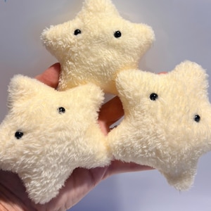 May include: Three small, pale yellow star-shaped plush toys made of a soft, fuzzy, textured fabric. Each star features two small, round, black bead-like eyes positioned on the front surface, giving them a simple and consistent appearance.