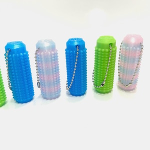 Puede incluir: Seis coloridos juguetes sensoriales cilíndricos con superficies texturizadas y cadenas de bolas adjuntas. Los juguetes son en tonos de verde, azul y un patrón tie-dye de rosa y azul. Están dispuestos sobre una superficie blanca.