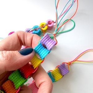 Op de afbeelding: Een kleurrijke armband met een reeks verbonden, getextureerde, rechthoekige kralen in verschillende kleuren, waaronder rood, oranje, geel, groen, blauw en paars. De armband is geregen op elastische koorden.